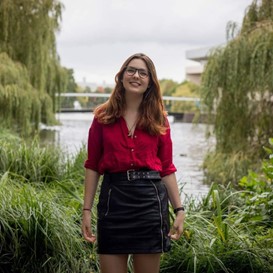 A photo of Steph stood in front of a large lake, with greenery on either side. She is a white woman, in a red shirt and black skirt, with brown hair and glasses, and she is smiling at the camera.