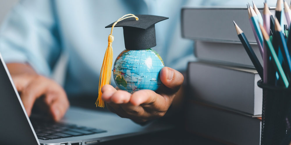 Hand holding miniature globe with graduation cap on top, to represent world higher education